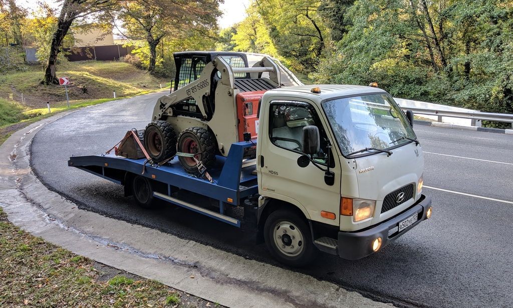 A white flatbed truck transporting a small construction vehicle on a winding road surrounded by trees.
