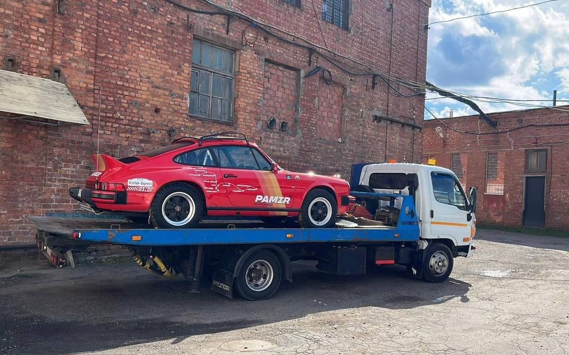 Red sports car on a blue tow truck in front of a brick building under a cloudy sky.