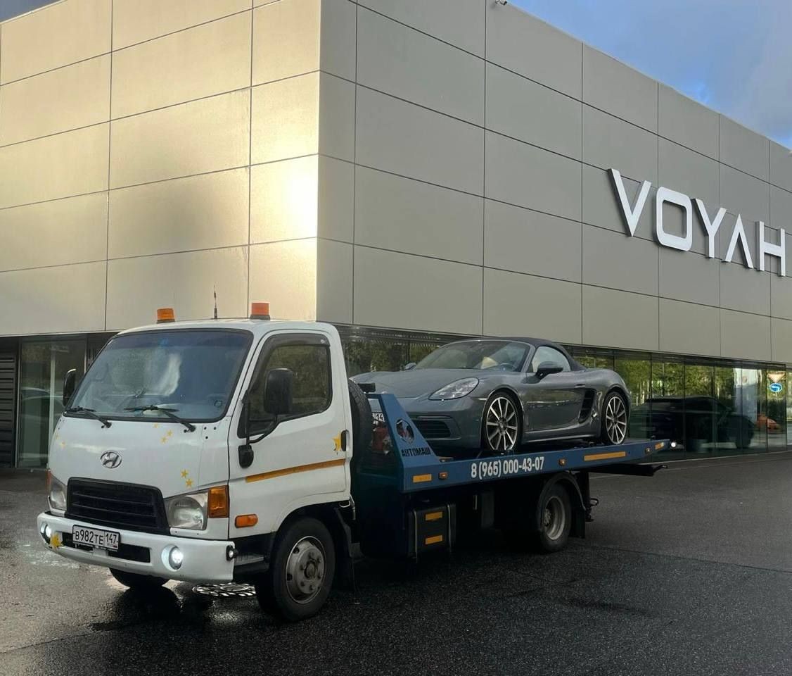 Flatbed tow truck carrying a sports car parked in front of a modern auto dealership.